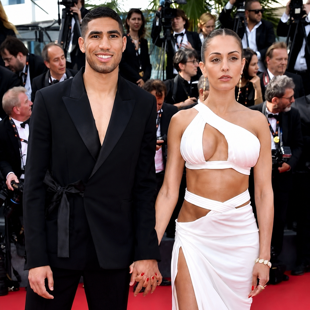 Achraf Hakimi in a black tuxedo holding hands with a woman in a white cutout dress at a red carpet film festival event