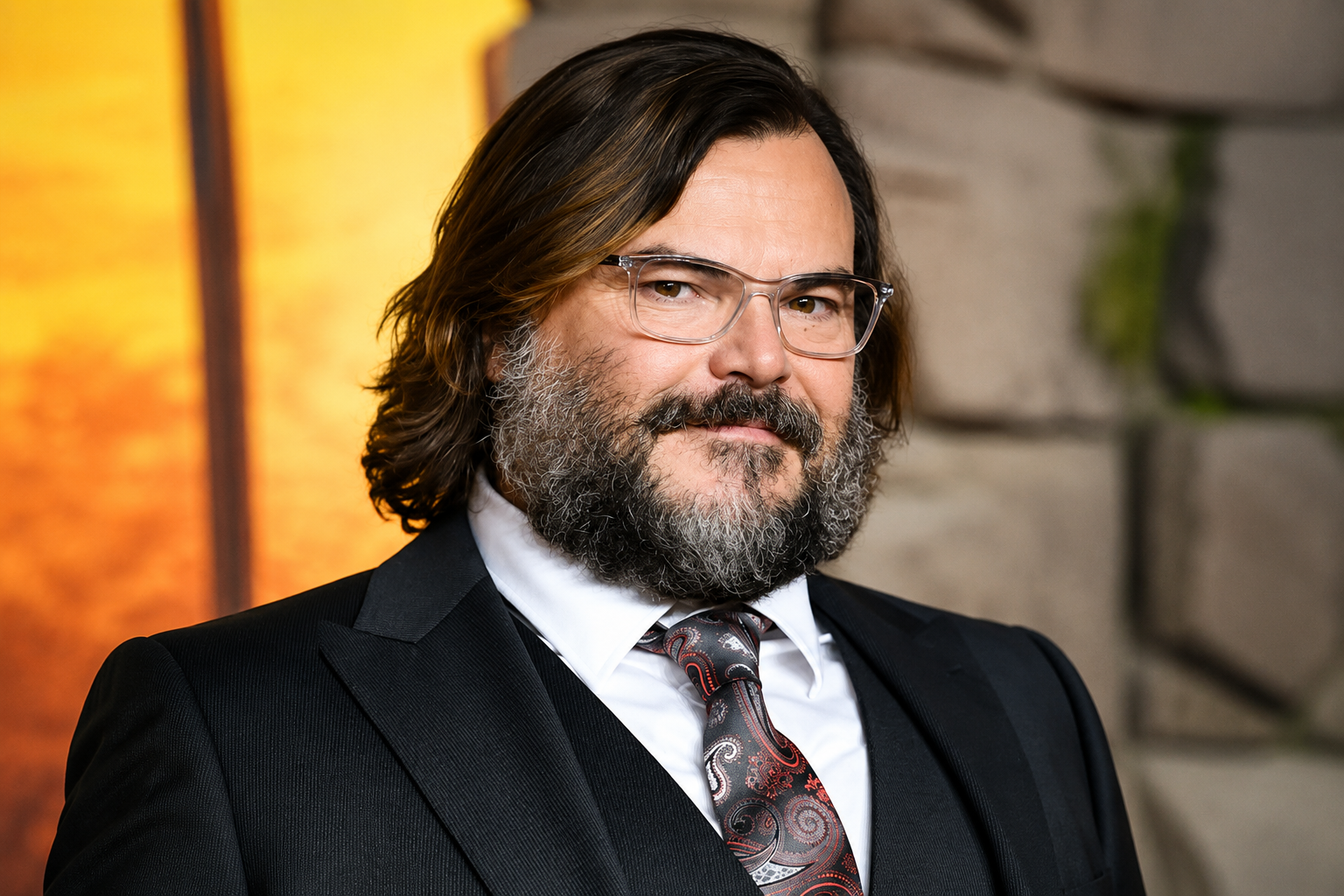 Jack Black net worth 2026 – wearing a black suit, white shirt, and paisley tie in a formal portrait