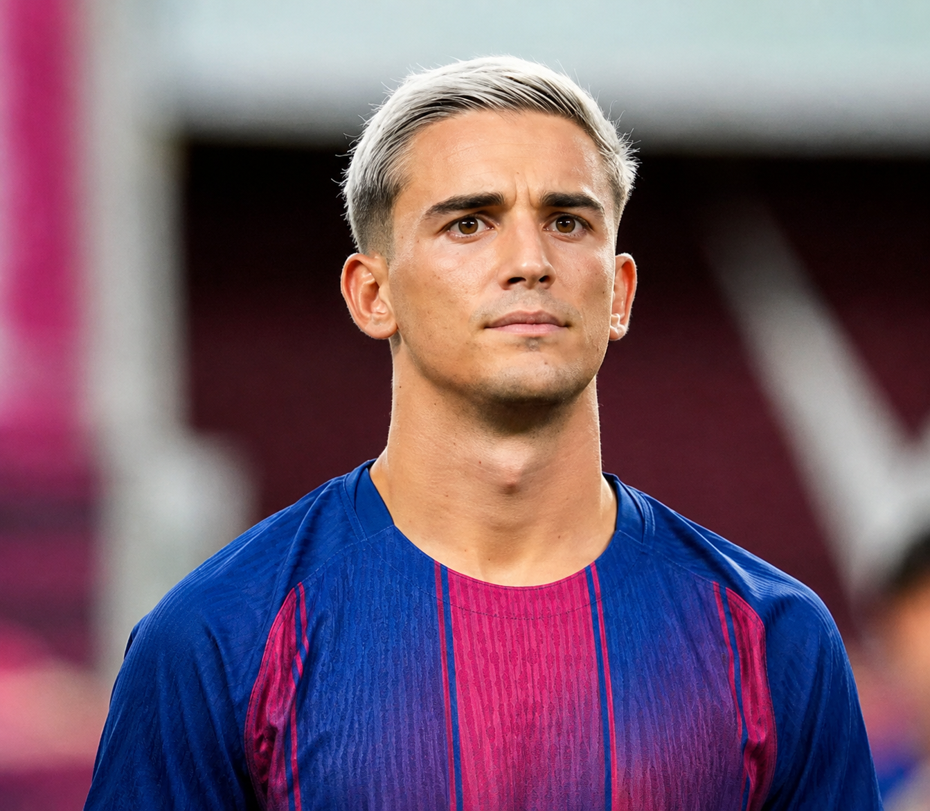 Young football player with short silver-blond hair wearing a blue and red jersey, posing with a focused expression in a stadium.