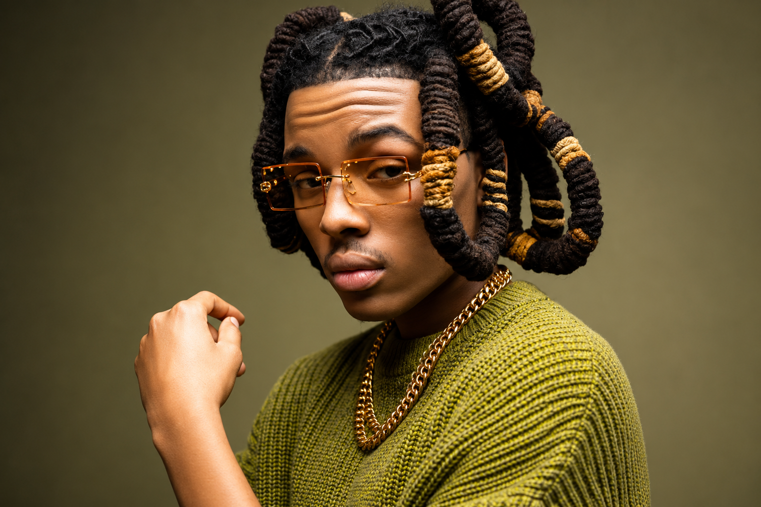 Young man with locs, gold glasses, and green sweater in a studio portrait