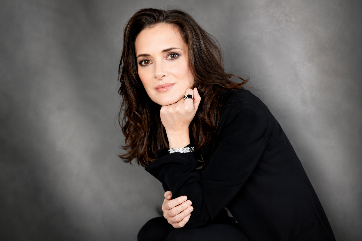 Winona Ryder posing in a black outfit against a gray studio backdrop
