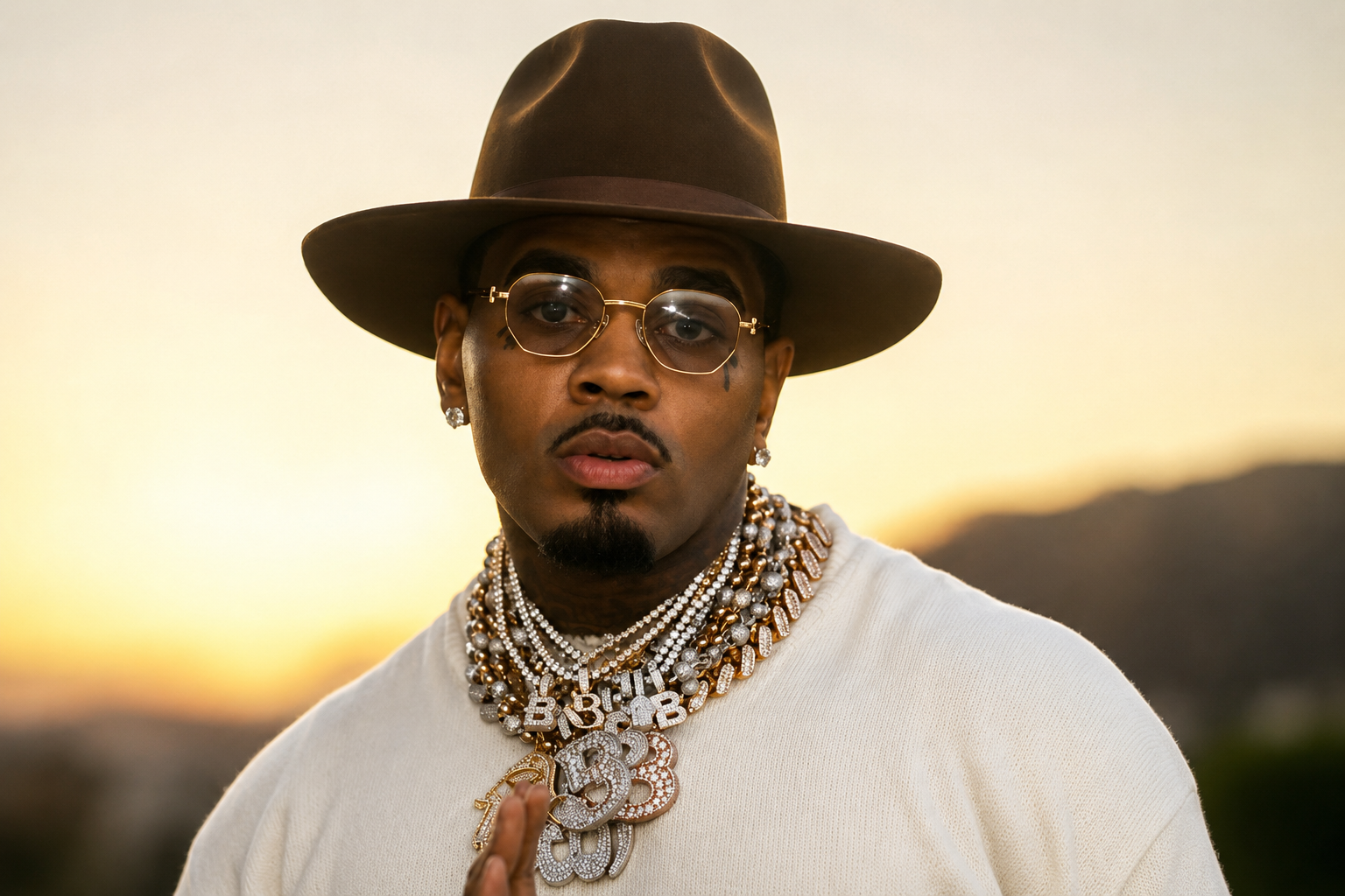 Kevin Gates wearing a brown hat, glasses, and layered diamond chains during an outdoor portrait