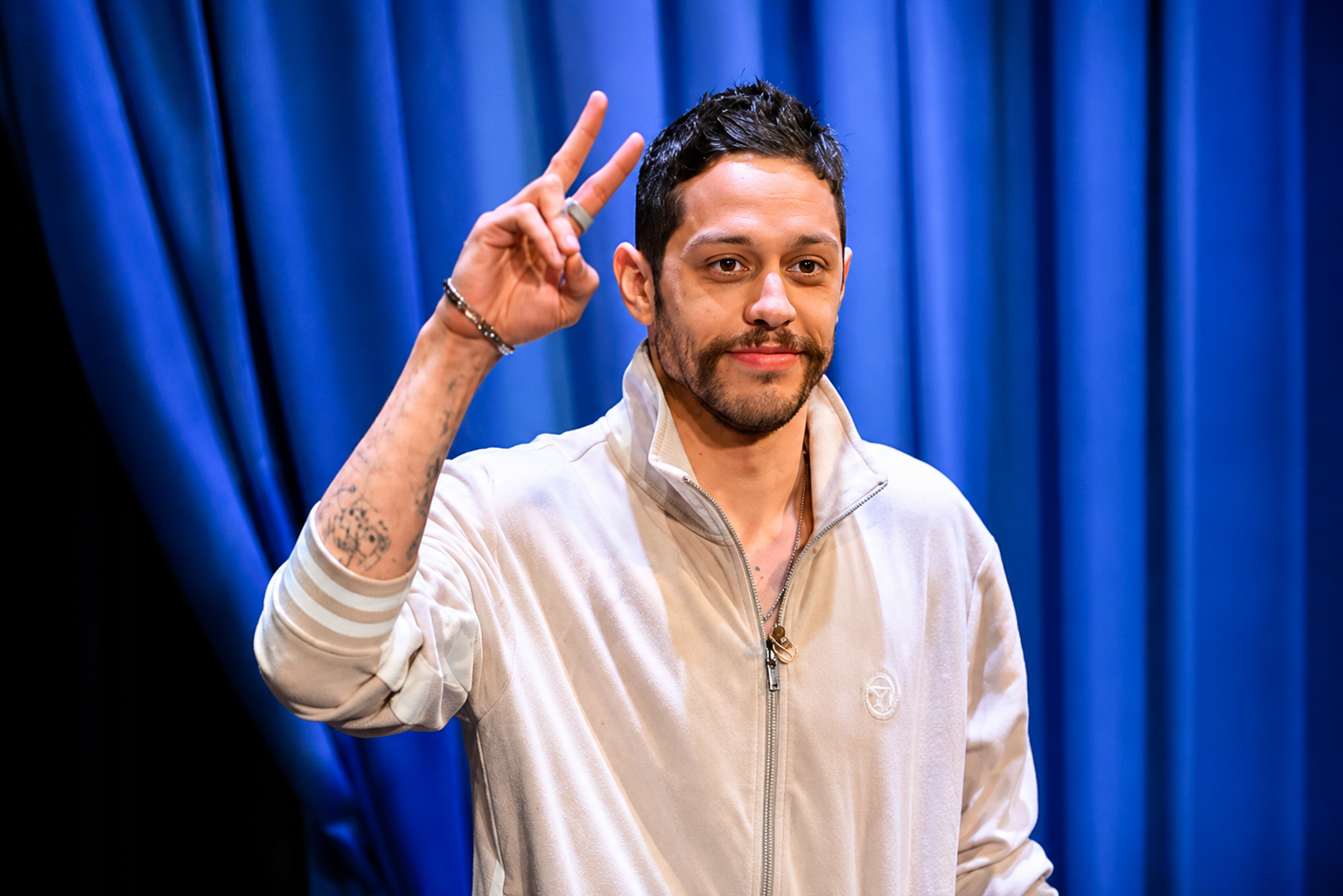 Pete Davidson making a peace sign while wearing a light-colored jacket against a blue background