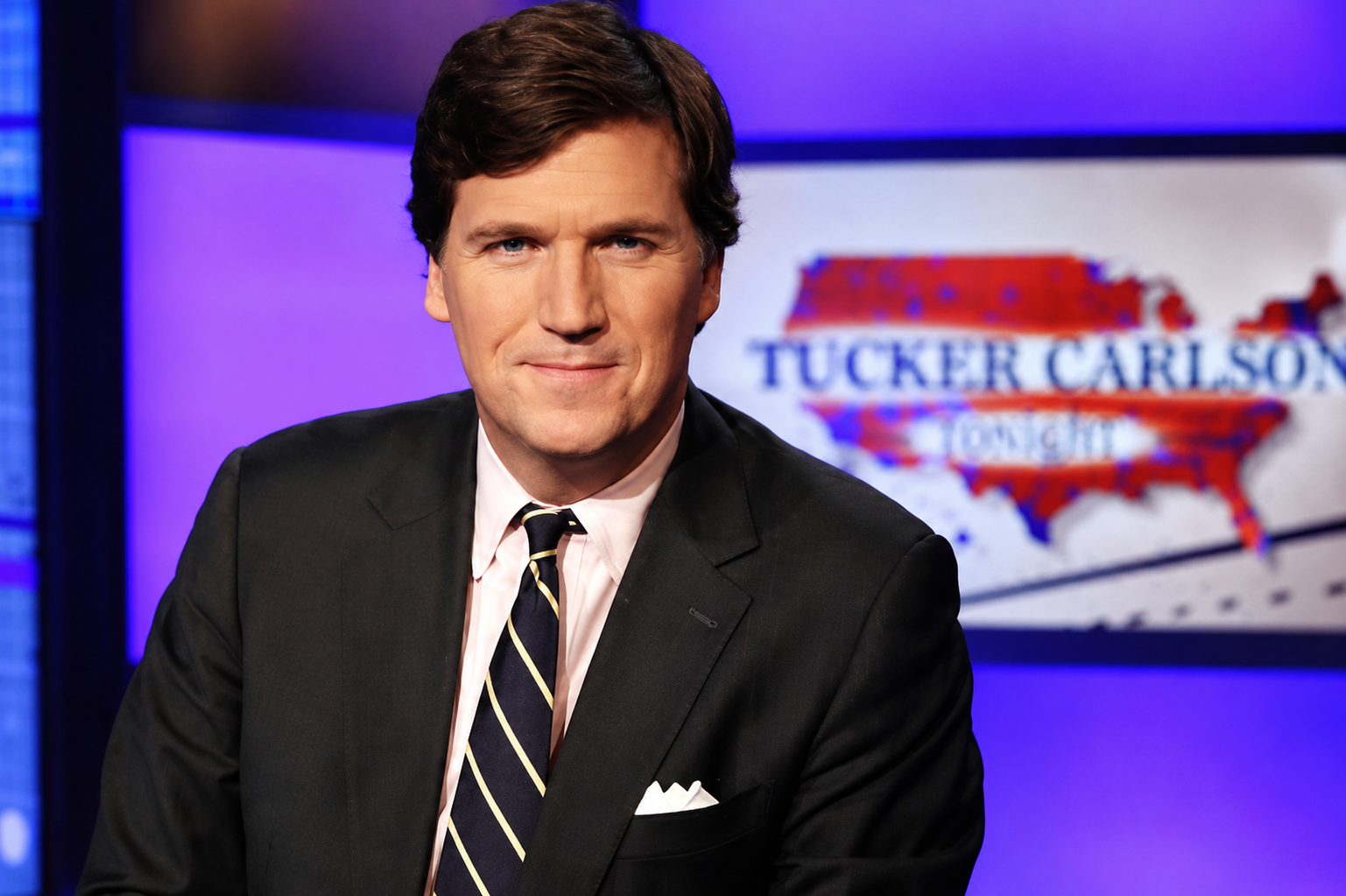 Tucker Carlson sitting in a television studio wearing a black suit and striped tie