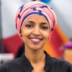 Ilhan Omar net worth 2026 - smiling portrait in a pink and navy headwrap and black top at an outdoor event