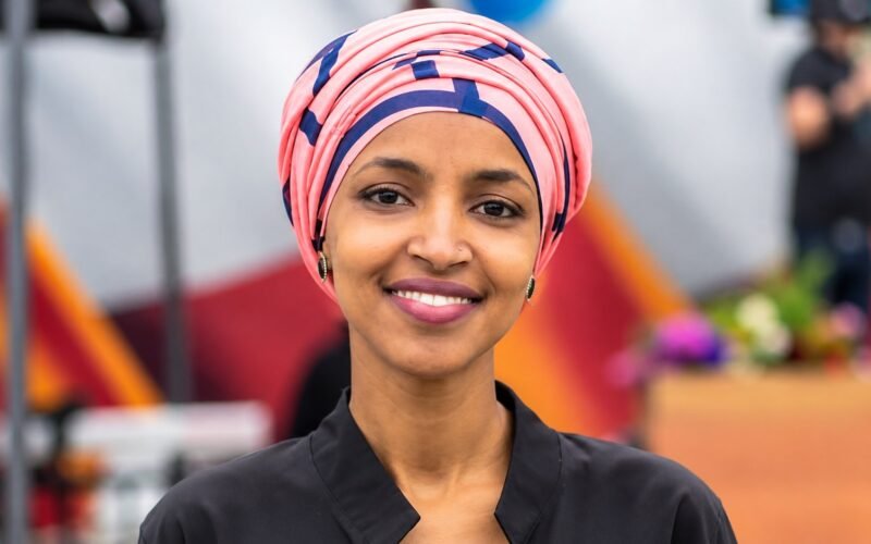 Ilhan Omar net worth 2026 - smiling portrait in a pink and navy headwrap and black top at an outdoor event