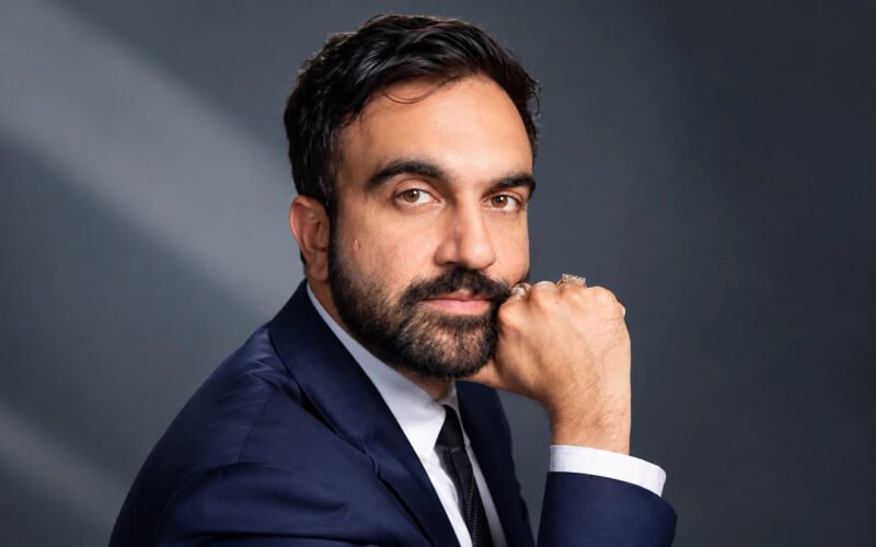Zohran Mamdani posing in a navy suit for a formal portrait
