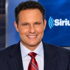 Brian Kilmeade smiling in a pinstripe suit and red tie in a SiriusXM studio