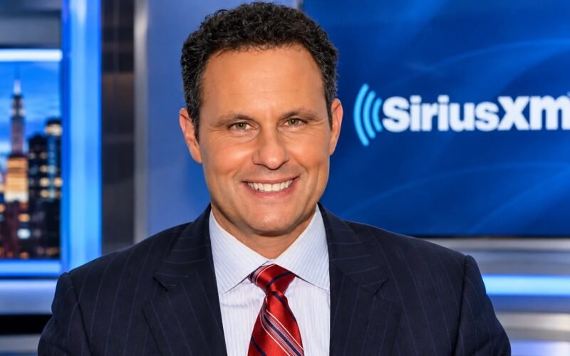Brian Kilmeade smiling in a pinstripe suit and red tie in a SiriusXM studio