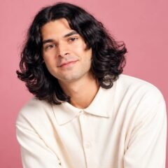 Daniel Zolghadri with long dark wavy hair in a cream shirt against a pink background