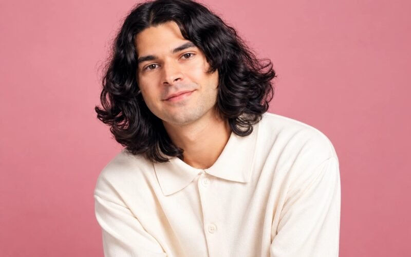 Daniel Zolghadri with long dark wavy hair in a cream shirt against a pink background